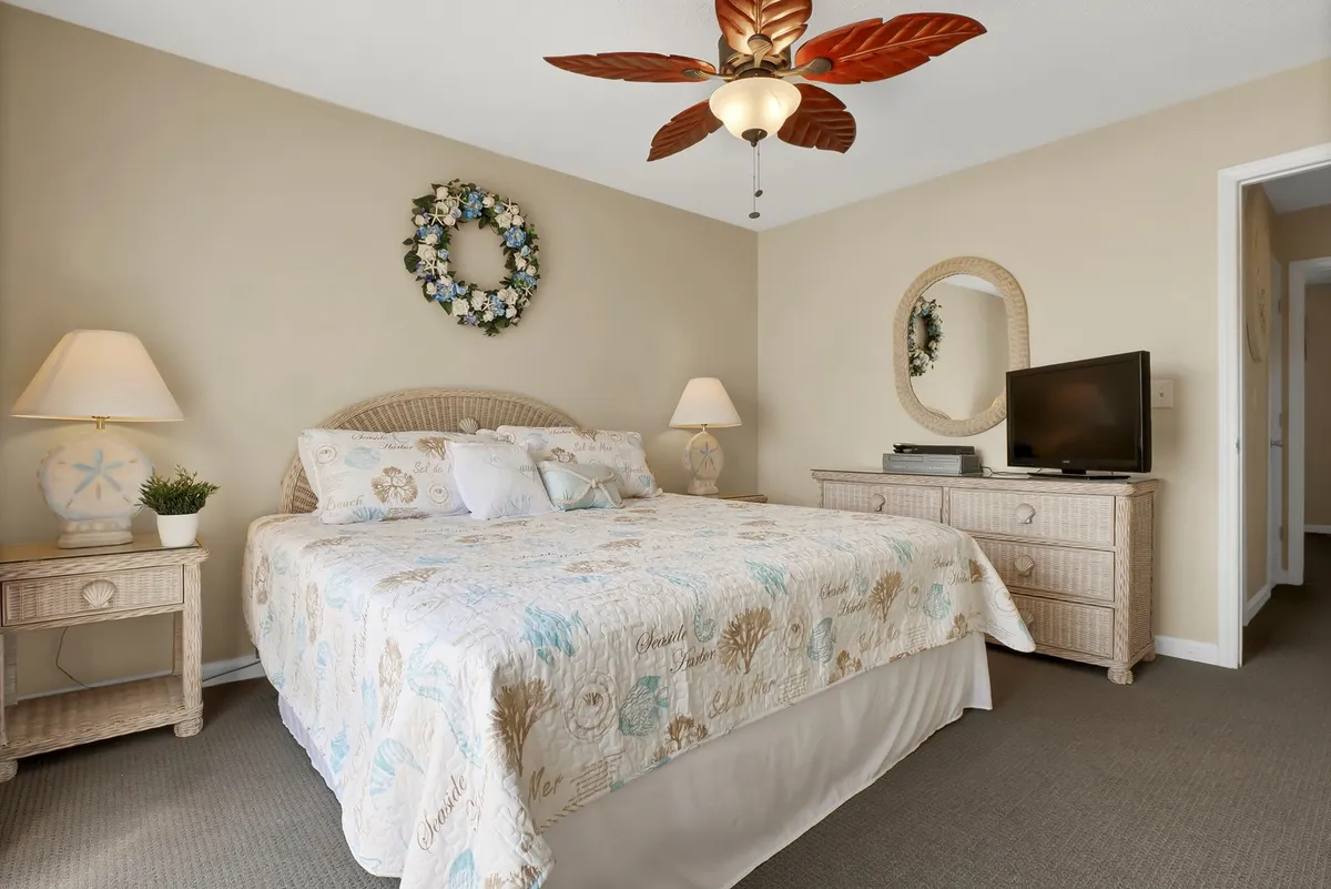 A serene bedroom with a bed featuring decorative bedding, a ceiling fan with a leaf design, a wall wreath, a nightstand with a lamp, and a television mounted on the dresser. The room exudes tranquility and includes details that suggest comfort and privacy for personal relaxation.