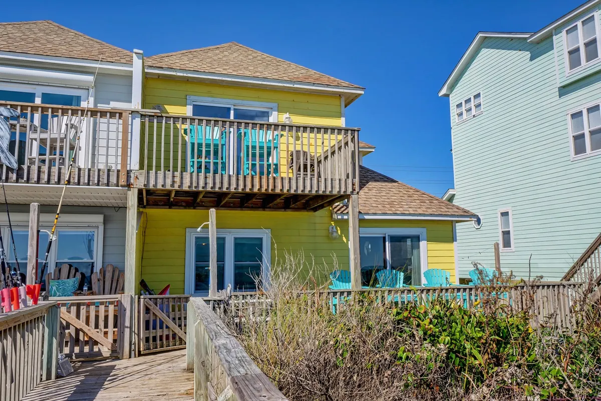 The yellow house exterior with a welcoming deck and blue chairs on the balcony overlooks a boardwalk stretching towards the beach. The lively architecture and playful seating hint at both relaxation and social connection with the ocean just beyond.