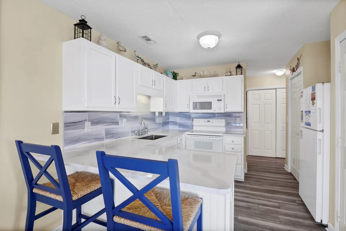 A modern, newly renovated kitchen with bright blue bar stools, white cabinetry, a marble-like backsplash, and a central island—ideal for casual meals, cooking, and socializing. The cheerful ambiance suggests a space meant for gathering and preparing family feasts.