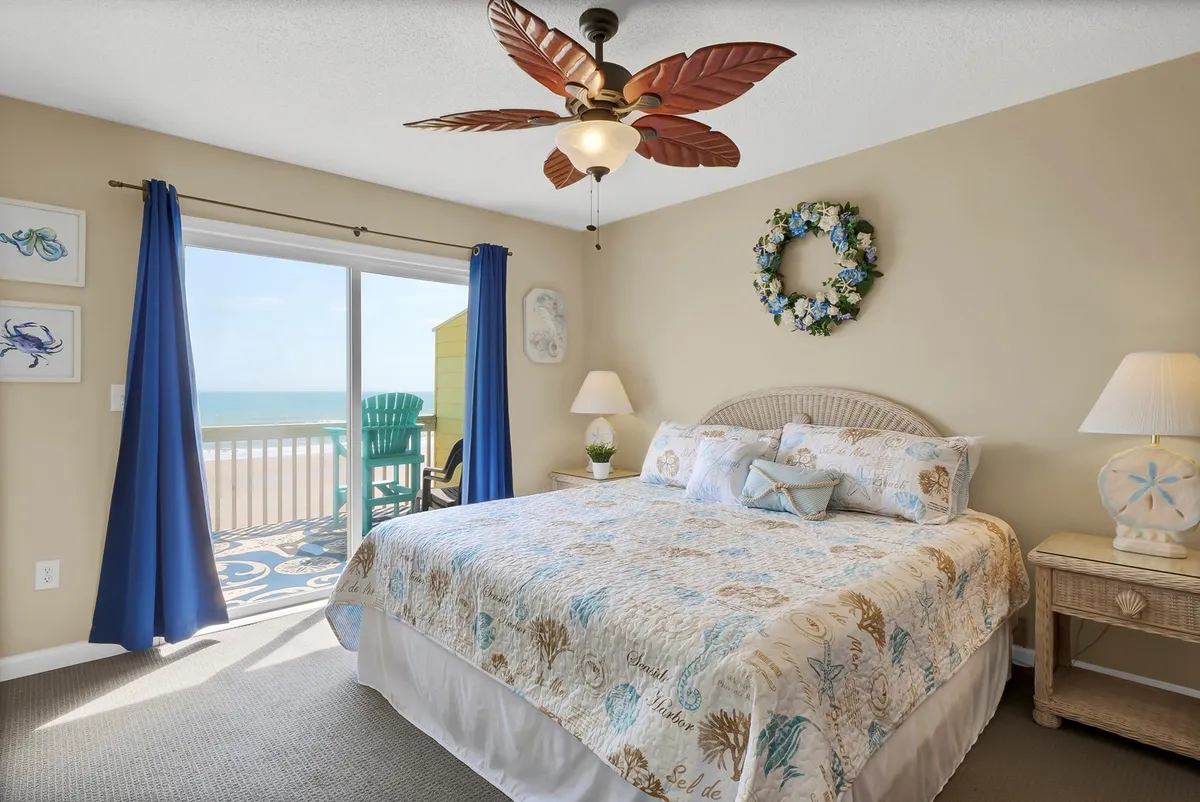 A spacious bedroom featuring a large bed with decorative bedding is at the center, accented by a ceiling fan with tropical flair. A sliding glass door leads out to a private balcony, and colorful curtains add vibrancy, while wall art provides a homely touch. Ample natural light fills the room, and the layout suggests both comfort and a hint of luxurious retreat.