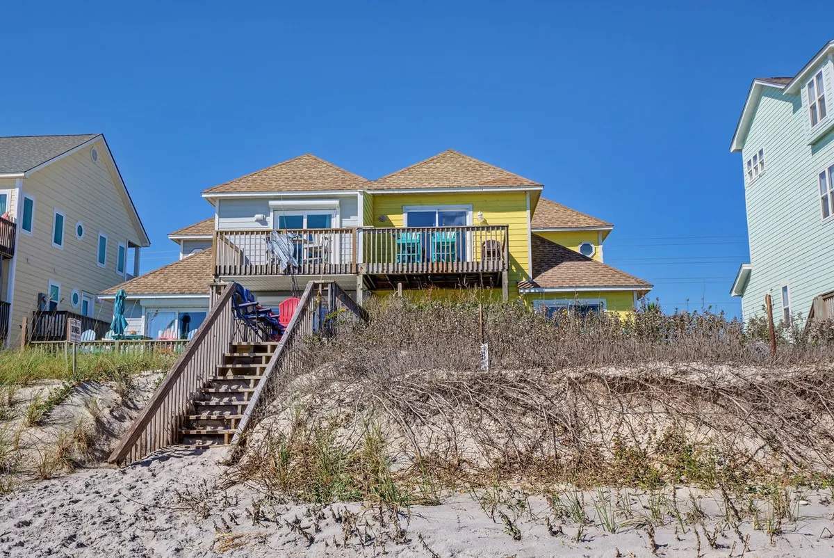A vibrant, colorful house with a spacious wooden deck and a staircase that leads directly down to the sandy beach. The scene feels inviting and cheerful, highlighting easy access from the house to the shoreline and plenty of outdoor space for gatherings or play.