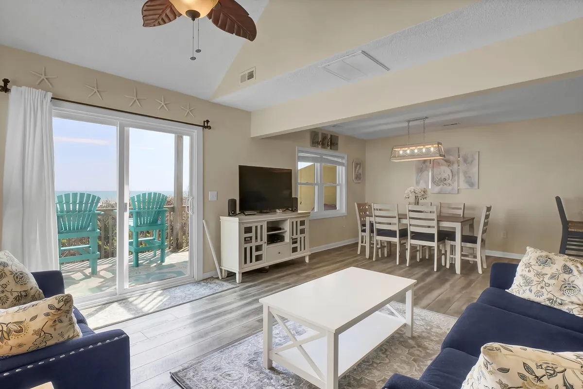 A cozy living room featuring large windows overlooking the ocean, bright blue deck chairs, a stylish coffee table, and a comfortable sofa with decorative pillows. The setting exudes a family-friendly atmosphere for evening games or relaxed gatherings.