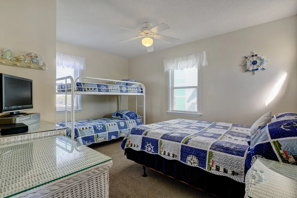 A cheerful, family-oriented bedroom containing a bunk bed and a double bed, both dressed in decorative quilts. A ceiling fan overhead and sunlight streaming through the window contribute to the cozy, playful atmosphere. The space feels perfect for children or families sharing a room.