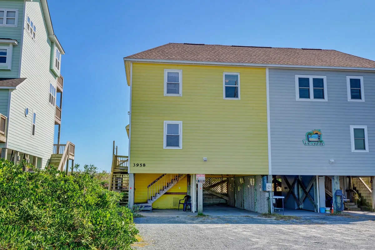 A cheerful exterior shot of the yellow duplex house with a staircase leading up and the house number '3958' visible. The inviting porch offers glimpses of the ocean beyond, with the home's architecture clearly indicating the duplex setup ideal for group stays.