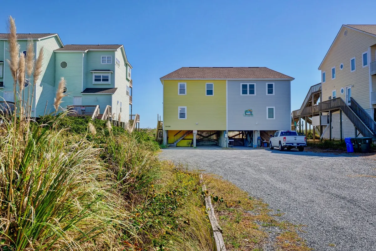 This image captures the exterior of a cheerful yellow house surrounded by greenery, with a gravel driveway in the foreground. The scene conveys a welcoming, family-friendly retreat perfect for gatherings, outdoor games, and sunshine-filled relaxation.