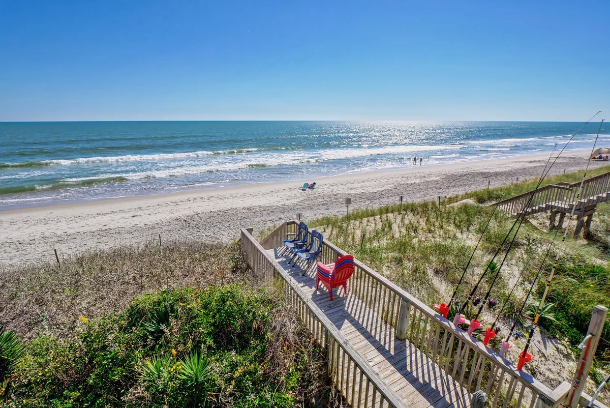 A tranquil wooden deck outfitted with colorful chairs overlooking the ocean, with fishing rods hinting at recreational activities and a sandy beach just beyond. The deck is spacious enough for several people to gather and relax together.