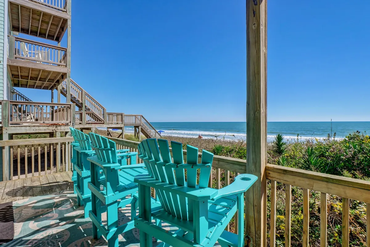 The image shows a spacious wooden deck overlooking the ocean, with vibrant turquoise chairs arranged for relaxation and conversation. The sun rises over the sparkling water, bathing the scene in a warm, golden light, creating an inviting and tranquil atmosphere ideal for gatherings.