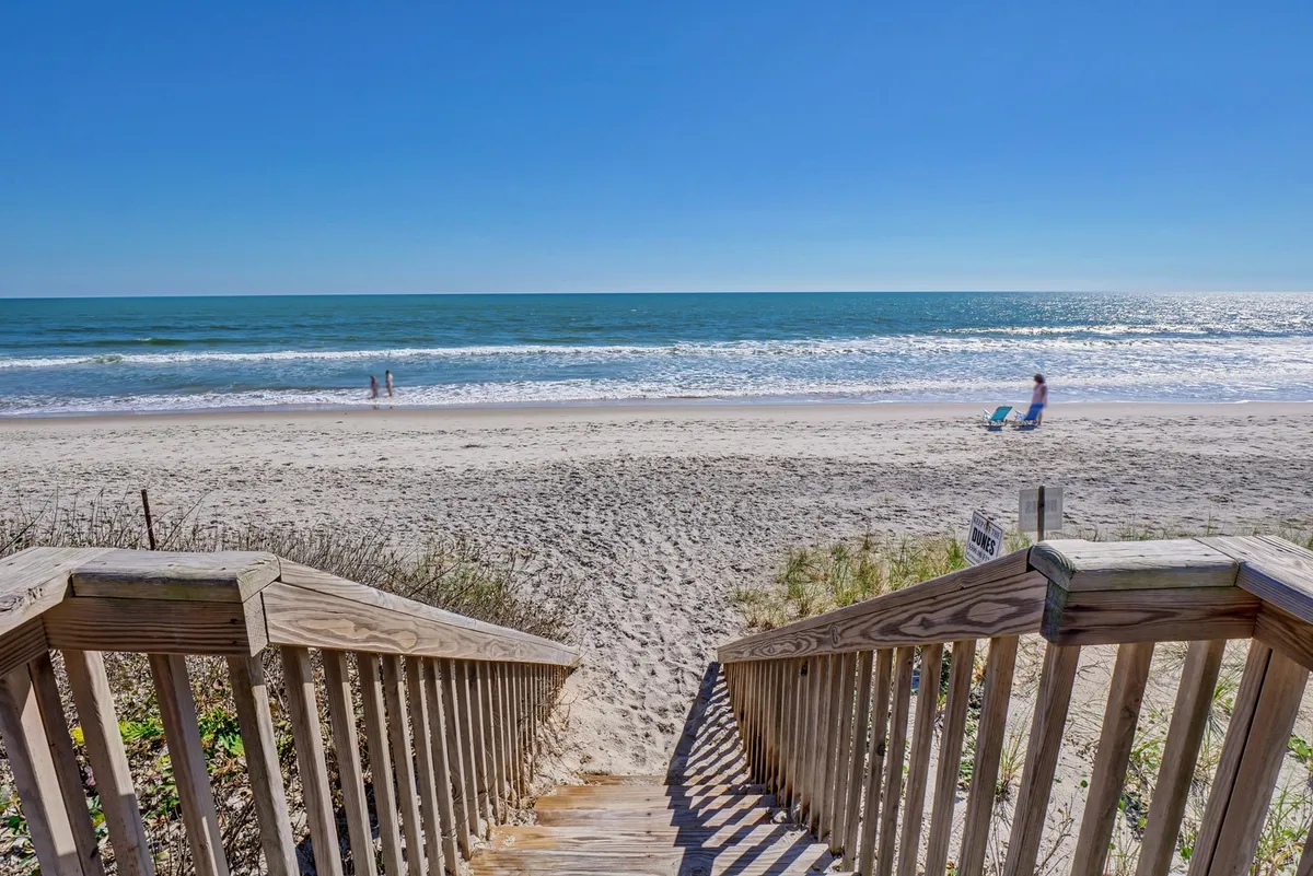 Wooden steps lead down to a sandy beach, with gentle ocean waves and people enjoying the shoreline. The scene effortlessly connects the home to the beach, showing just how close the sand and surf are to the property.