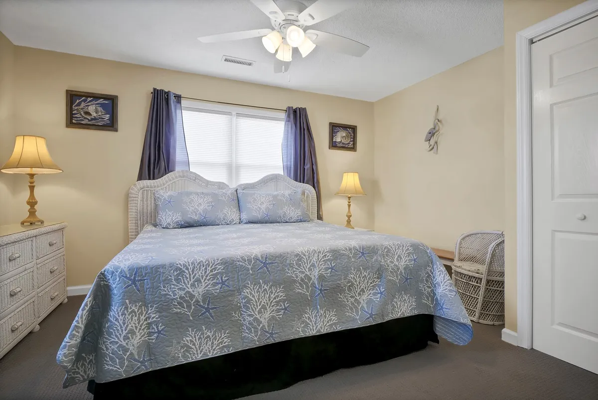 A serene bedroom with a bed dressed in blue coral-patterned bedding, two bedside lamps, and a window with sheer curtains that allow gentle natural light to fill the space. The decor is peaceful and inviting, signaling rest and relaxation within a private retreat.
