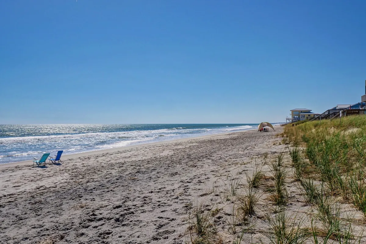 A tranquil stretch of sandy beach bordered by gentle dune grass, with lounge chairs and a small tent set up for beachgoers. The blue ocean extends to the horizon under a clear sky, evoking the quintessential beachfront family experience.