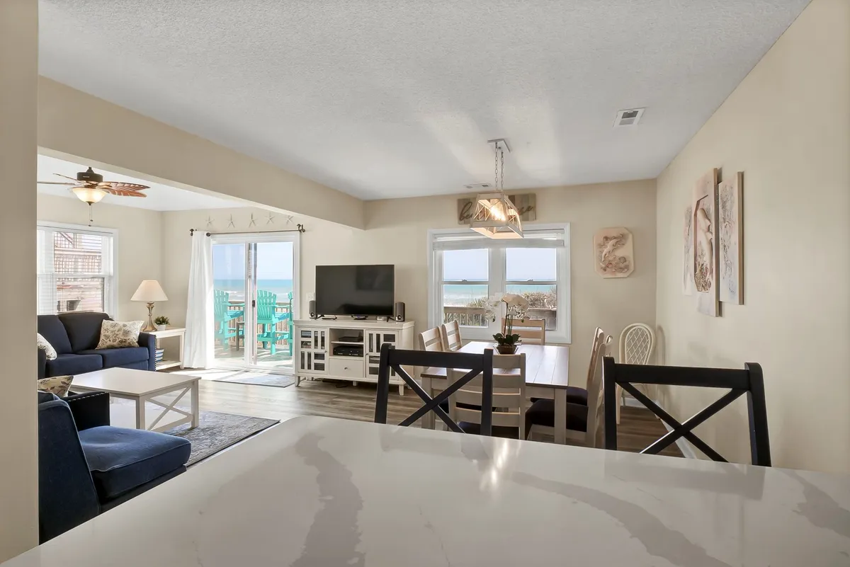 A bright, open living area featuring comfortable seating, a large dining table, a TV unit, and sliding glass doors that lead directly to a deck. Natural light fills the space, and the room invites group gatherings, meals, and relaxation. The deck with ocean views is visible through the glass doors.